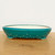 A side view of a 12-inch oval Yixing ceramic bonsai pot (item No. 2512A), showcasing its vibrant teal glazed finish and distinctive riveted design. The shallow pot features a line of raised decorative dots around its lower exterior and rests on four subtle feet. Its interior is a pale cream color, providing a clean contrast to the turquoise exterior. The pot is positioned on a light-toned wooden surface against a plain white backdrop, highlighting its elegant shape and textured glaze.