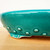 A detailed close-up shot showcasing the side and base of a 12-inch oval Yixing ceramic bonsai pot. The pot is finished in a vibrant, glossy turquoise glaze that features a dense, intricate crackle pattern throughout. A series of raised, rounded rivets are arranged horizontally along the lower portion of the pot's side, adding a decorative textural element. The image also highlights one of the pot's cloud-style feet, revealing the light tan, unglazed ceramic beneath the thick teal glaze. The pot is positioned on a light-colored wooden table against a plain white background, emphasizing its rich color and artisanal details.