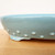 A close-up, low-angle shot captures the side and base of a 12-inch oval Yixing ceramic bonsai pot. The pot is finished in a glossy, pale sky-blue glaze that features subtle, natural dark speckles throughout. Decorating the lower curve of the pot is a series of raised, rounded "rivet" or stud-like accents that add a traditional, textured detail. The pot rests on small, integrated feet with simple carved vertical lines, shown sitting on a light-colored wooden surface against a clean, white background.