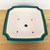 A top-down view of a 9-inch square Yixing ceramic bonsai pot with a deep teal-green glaze on its outer rim and sides. The interior of the pot is a smooth, unglazed cream color, featuring one large central drainage hole and four smaller holes positioned toward the corners for securing a tree with bonsai wire. The pot has an elegant shape with slightly curved sides and recessed corners, resting on a light-colored wooden surface against a neutral white background.