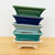 A vertical stack of five small, 4.5-inch glazed Yixing ceramic bonsai pots in various shades. Each rectangular pot features a flared rim, subtle decorative indentations around the center, and four small feet at the base. From top to bottom, the glossy colors include forest green, dark navy blue, powder blue, teal green, and off-white. The pots are displayed on a light-colored wooden surface against a plain white background, highlighting their smooth finish and classic design.