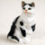 A hand-painted black and white shorthaired tabby cat figurine sitting on a light-colored surface. This decorative miniature cat, often used as a bonsai tree accessory or fairy garden ornament, features a predominantly white body with striking black patches on its head, ears, back, and tail. The cat has a curious expression, with vibrant green eyes looking slightly up and to the side, and a soft pinkish-orange nose. The detailed sculpting realistically mimics the texture of short cat fur, making it a charming addition to miniature landscapes or home decor.