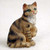 A detailed, hand-painted miniature brown shorthaired tabby cat figurine, ideal for a bonsai display or miniature garden. The figurine depicts a sitting cat with realistic proportions, featuring a warm tan coat accented with dark brown tabby stripes and a white bib area on its chest. Its head is turned slightly to the side with alert green eyes and a small pink nose. One of its front paws is delicately lifted in a playful gesture. The cat sits on a plain, light-colored textured surface, highlighting the fine craftsmanship and textured fur detail of the collectible piece.