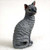 A detailed miniature figurine of a Blue Cornish Rex cat sits gracefully in an upright, alert pose against a soft white background. The feline sculpture is rendered in a deep slate grey color, characteristic of the "blue" coat variety, and features the breed's signature tight, wavy texture across its entire body. Its slender, athletic form is captured as it sits on its haunches with a long tail curled neatly around its base. The figurine's face is turned slightly to the side, showcasing large, pointed ears and striking green eyes that provide a lifelike, inquisitive expression. This collectible cat statue is designed as a decorative accessory, ideal for adding a touch of personality to a bonsai garden display or a desktop collection.