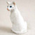 A small, detailed white Oriental Shorthair cat figurine is captured in a close-up, three-quarter view against a neutral, off-white background. The figurine depicts the cat in an upright seated position, with its head turned slightly to the left. 

The cat’s features include striking, almond-shaped bright blue eyes and a small, delicate nose and mouth painted in a soft pinkish-orange hue. The figurine's body has a realistic, etched texture that mimics the appearance of short, fine fur. The craftsmanship suggests a hand-painted finish, with subtle shadows highlighting the lean, elegant musculature characteristic of the Oriental Shorthair breed. This miniature cat figurine is designed as a decorative accessory, often used to add character and a sense of scale to bonsai tree displays or fairy gardens.