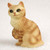 A detailed miniature figurine of a red tabby Manx cat, captured in a sitting position against a plain, light-textured background. The figurine is meticulously crafted with a realistic fur-like texture across its body. Its coat is a warm ginger-orange with darker reddish-orange tabby stripes visible on its side and front leg, while its chest and muzzle are a lighter cream color. The cat's face features large, dark, expressive eyes, a small pinkish-red nose, and a subtle, pleasant smile. One of its front paws is raised slightly toward its chest, as if in a grooming gesture. Characteristically for a Manx breed, the figurine shows a rounded rear end without a long tail. This decorative cat statue is designed as a charming accent for bonsai tree displays or miniature fairy gardens.