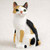 A detailed miniature figurine of a Japanese Bobtail cat, featuring a classic "tortoise and white" (calico) coat pattern. The cat is depicted in a sitting, alert pose against a soft, neutral off-white background. Its primarily white body is decorated with distinct patches of ginger-orange and black across its back, head, and legs. 

The figurine captures the breed's unique characteristics, including its signature short, "bobbed" tail, large attentive green eyes, and small pink nose. The surface of the figurine is finely textured to mimic the appearance of real fur. This charming miniature is designed as a decorative accessory, perfect for adding a touch of personality to a bonsai tree display or a tabletop Zen garden.