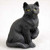 A detailed miniature figurine of a black cat sitting in an upright position on a plain, light-colored background. The figurine features realistic, textured black fur and has its front right paw slightly raised, as if caught in mid-motion while grooming. It has striking, round yellow eyes with dark pupils, giving it a lifelike expression. This decorative shorthaired tabby figurine is designed as a small-scale accessory, perfectly sized for adding character to a bonsai tree display or a miniature fairy garden.