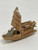 A high-resolution, close-up photograph of a miniature Chinese mudman figurine depicting a traditional skiff boat, designed as a decorative accessory for bonsai trees. The handcrafted ceramic boat is finished in a glossy, light brown or tan glaze. It features a tall, vertical sail with horizontal ribs, textured to resemble woven bamboo or fabric. 

At the bow of the skiff sits a small, unglazed grey mudman figure, hunched forward as if rowing or tending to the vessel. The middle section of the boat carries several thick, horizontal log-like structures, some of which are accented with patches of deep reddish-purple glaze. A section of the boat's side features a cross-hatched, lattice-like pattern. The entire figurine is set against a clean, neutral white background, emphasizing the intricate textures and traditional craftsmanship of the piece.