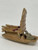 A detailed, side-profile view of a small, glazed Chinese mudman figurine depicting a traditional skiff boat. The ceramic figurine is finished in a warm, glossy tan glaze. At the bow of the boat sits a miniature mudman figure dressed in a muted grey-green robe, shown in a contemplative pose. Behind the figure rises a tall, rectangular sail with horizontal ridges, reminiscent of a traditional junk boat sail. The midsection of the skiff features a canopy structure made of three horizontal, bamboo-like poles, each decorated with dark reddish-purple spots. Beneath these poles, the sides of the boat display a fine, textured pattern mimicking a woven mat. The boat rests on a plain, light-colored surface, highlighting its handcrafted details and polished glaze. This miniature piece is a typical example of a decorative accessory used to add character and narrative to a bonsai tree display.