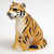 A detailed miniature tiger figurine is depicted in a sitting position against a plain white background, shown from a three-quarter angle. The figurine features a realistic orange-brown coat with distinct black vertical stripes and a white underbelly, chest, and muzzle. Its mouth is slightly agape, revealing teeth and a pink interior, as if in a soft growl. One front paw is lifted slightly, and its tail is curled around its back haunches. The surface of the figurine is finely textured to mimic the appearance of fur. This small animal figurine is designed as a decorative accent for bonsai tree arrangements, adding a touch of wildlife and character to miniature indoor garden displays.