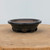 A detailed side-view photograph of a handmade ceramic bonsai pot, identified as Brandon Besserer No. 10. This round, shallow pot features a wide, flared rim and sits on several small, chunky feet that provide elevation. The exterior is coated in a dark, metallic charcoal glaze with a subtle, mottled texture, while the interior and the underside of the feet showcase the unglazed, reddish-brown natural clay. The pot is positioned in the center of a light-colored wooden display stand against a neutral, off-white background, highlighting its artisanal craftsmanship and earthy aesthetic.