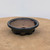 A handcrafted ceramic bonsai pot by artist Brandon Besserer (No. 10). This round, shallow pot features a wide, rounded rim and a dark, charcoal-metallic glaze that contrasts with the unglazed, speckled reddish-brown clay of the interior. The pot's exterior walls taper down to a series of decorative cloud-shaped feet at the base. Inside, a large central drainage hole and several smaller wire holes are visible. The pot is centered on a light tan, textured surface against a stark white background, highlighting its artisanal detail and unique finish.