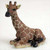 A detailed, miniature giraffe figurine designed as a decorative accessory for a bonsai tree or indoor fairy garden. The small giraffe is depicted in a peaceful, resting pose, sitting with its legs tucked neatly beneath its body. It features a realistic pattern of deep brown patches separated by a network of cream-colored, lattice-like lines across its body and long neck. The figurine captures the giraffe's characteristic features, including small horns (ossicones), large dark eyes, pointed ears, and a dark-colored snout. The textured surface of the figurine mimics animal hair, providing a lifelike appearance for use as a charming accent piece in a miniature landscape or home decor display.