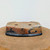 An upside-down view of a shallow, oval-shaped ceramic bonsai pot, showcasing its unique handcrafted details. This piece, created by American ceramicist Bruce Lenore, is made from a warm, earthy terracotta-colored clay with a rugged, pitted texture and small carved indentations. It features artistic accents of dark indigo-blue glaze that swirl across its sides and along the bottom rim. On the base of the pot, which faces upward in the image, two round drainage holes are visible, along with four integrated low-profile feet. The pot sits on a circular wooden platform, presented against a clean, off-white background to highlight its artisanal quality and rustic aesthetic.