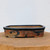 A side view of a rectangular, handmade ceramic bonsai pot crafted by American artist Bruce Lenore. The pot features rounded corners and a slightly flared rim finished with a dark, glossy glaze. Its body displays a rustic, earthy texture with numerous small triangular indentations and a color palette of warm orange and sandy tan. Abstract dark blue or black markings, resembling expressive brushstrokes, are layered over the textured surface. The pot is supported by small, integrated rectangular feet and rests on a natural wood surface against a minimalist white background.