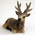 A small, detailed figurine of an elk bull lying in a resting position, designed as a decorative accessory for a bonsai tree or miniature garden. The figurine is crafted with realistic textures, featuring multi-pointed antlers and a coat of earthy brown shades that transition from light tan on the face and neck to a darker chocolate brown along the back. The elk's head is turned slightly toward the viewer, showcasing small, glossy black eyes and a prominent black nose. Its black hooves are tucked neatly beneath its body, and its ears are perked up. The figurine is set against a plain, soft-white background, highlighting its intricate design and suitability for adding a touch of wildlife to a natural bonsai landscape display.