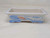 A side-view photograph of a 7-inch rectangular Yixing bonsai pot, showcasing its hand-painted details. The pot is made of white ceramic with a smooth glaze. The front panel features a serene, traditional landscape scene painted primarily in blue, with a small house in the center accented by a reddish-orange roof. The landscape includes textured hills, distant mountains, and tiny birds in flight, all contained within a simple blue frame. A matching blue geometric pattern lines the flat top rim, and the four short, decorative feet at the base are adorned with a blue cross-hatch design. The pot sits on a plain, light-colored surface, emphasizing its elegant and traditional design.