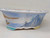 An eye-level, side view of a 10-inch hand-painted Yixing ceramic bonsai pot, showcasing a traditional landscape scene. The pot has an elegant oval shape that tapers toward a footed base and features a wide, flared rim. 

The exterior is decorated with a serene, hand-painted watercolor-style scene against a pale blue background. On the right side, large, craggy mountain peaks are rendered in shades of blue and gray with black ink-like outlines. Sparse green pine trees grow along the mountainside. To the left, a small brown sailboat with tiny figures on board glides across a body of water, with a soft brown shoreline visible in the distance. A flock of small black birds in 'V' formations flies across the light sky. 

The white glaze of the pot has a subtle crackle finish, adding a sense of antiquity. The top rim is accented with a delicate pattern of blue scrolls and small brown dots. This decorative Yixing bonsai pot combines functional design with fine artistic detail, ideal for displaying a mature bonsai tree.