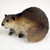 A detailed miniature figurine of a beaver, designed as a decorative accessory for bonsai trees or fairy gardens. The figurine is captured in a crouched position, facing left against a plain white background. It features realistic textures, with dark brown, coarse-looking fur along its back that transitions to a lighter sandy-tan on its belly and face. A prominent, flat, paddle-shaped tail with a bumpy, scale-like pattern is tucked alongside its body. Small details like tiny black eyes, dark ears, and a visible white front tooth add to its lifelike appearance. This small resin animal is crafted to bring a touch of wildlife and storytelling to miniature landscapes or penjing displays.