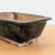 A high-angle, three-quarter view of a rectangular, handmade bonsai pot crafted by American artist Bruce Lenore. The ceramic pot features a glossy, dark espresso-to-black glaze that covers the exterior sides and the slightly overhanging, flat rim. The glaze has a rustic, artisanal quality with subtle drips and variations in depth. The lower edge and four small, block-shaped feet are left unglazed, revealing the natural, light-tan color of the clay body, with some white kiln wash visible. The interior of the pot is also unglazed, showing the raw material. The piece is resting on a light-colored wooden surface against a plain white background, highlighting the earthy tones and handcrafted texture of the vessel.