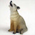 A small, realistic coyote figurine designed as a miniature bonsai tree accessory. The coyote is shown sitting on its haunches with its head tilted sharply back and its mouth wide open in a howl. The figure is intricately detailed with textured fur in shades of sandy tan and light brown, featuring darker grey accents along its back and on the tips of its ears and paws. This miniature howling coyote figurine is meant to add a sense of wilderness and scale when placed at the base of a bonsai tree or within a small-scale garden landscape.
