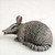 An intricate miniature armadillo bonsai tree figurine, shown in a side profile against a plain, light-colored background. This detailed sculpture is designed as a bonsai pot accessory and features a realistic, textured finish that resembles aged bronze or stone. The armadillo is depicted in a low, crouching pose with its long snout slightly open, prominent upright ears, and a thick, segmented tail curled along its side. Its protective armor is rendered with fine detail, showing characteristic horizontal bands across its back and a pebbled skin texture on its head and sides, making it a charming and characterful addition to a miniature landscape or bonsai arrangement.
