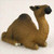 A detailed figurine of a dromedary camel, designed as a decorative accessory for a bonsai tree or miniature landscape. The camel is depicted in a restful, kneeling position on a white, textured surface that resembles fine desert sand. Crafted with realistic details, the figurine features a single hump and is painted in shades of warm medium brown, with darker brown accents on the top of its hump and head to mimic the texture of thick fur. Its long, elegant neck curves upward, leading to a small face with dark eyes and a blackish-brown snout. Subtle white highlights at the base represent its tucked-under feet. This charming dromedary camel miniature is an ideal addition for creating a desert-themed scene within a bonsai display, terrarium, or fairy garden.