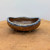 A small, oval-shaped ceramic bonsai pot, an American handmade piece by artist Bruce Lenore. The pot features a textured, earthy brown stoneware body with hand-carved lines and a spiral motif that mimics the natural appearance of wood grain or tree bark. Its irregular, wavy rim is finished with a thick, glossy blue-gray glaze that flows slightly over the edges, creating a striking contrast with the matte, rustic exterior. The vessel sits on small, stubby feet atop a weathered light brown wooden surface against a plain off-white background, emphasizing its organic and artisanal design.
