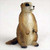 A detailed, hand-painted miniature figurine of a prairie dog, intended as a decorative accessory for a bonsai tree landscape. The small figure is depicted in a classic alert, upright posture, standing on its hind legs with its tiny front paws tucked against its chest. It features realistic fur textures in various shades of tan and warm brown, with small, glossy black eyes and a dark nose. Its short, rounded tail rests on the ground behind it. The figurine is set against a plain, off-white, slightly textured background, highlighting its intricate details and life-like appearance.