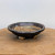 A high-angle, eye-level shot captures an American handmade ceramic bonsai pot by artist Bruce Lenore. The pot is a shallow, circular dish with a wide, slightly inward-sloping rim. It features a rustic, wabi-sabi aesthetic with a dark, metallic charcoal glaze that has a subtle iridescent sheen. In areas, the glaze thins to reveal the light, sandy-brown natural clay body, particularly on the interior and parts of the textured rim. The bonsai container sits on three small, sturdy, unglazed feet that show the raw tan clay. The pot is displayed on a light-colored, circular wooden slab against a neutral, off-white wall, emphasizing its handcrafted details and earthy tones.