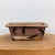 A rectangular, American handmade ceramic bonsai pot by artist Bruce Lenore is showcased against a plain white background. The pot features a muted, dusty rose or mauve glaze as its primary color, which is adorned with bold, expressive dark brown brushstrokes that wrap around the sides. Near the base, there are subtle hints of white and cream-colored glazes, adding depth to the finish. The pot is designed with a flared rim and small, integrated block feet at the corners. The interior of the pot reveals the natural, unglazed texture of the fired clay. This unique, artisanal piece is positioned on a rustic, circular light-wood base, highlighting its handcrafted character and earthy tones.