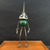 A whimsical, handcrafted robot sculpture stands centered on a polished wooden surface against a solid matte black background. This unique piece of found-object art is constructed from a variety of vintage materials. 

The robot’s torso is a weathered, dark green vintage croquet ball with visible scuffs that hint at its past life. Atop the body sits a head made from a silver vintage oil can lid, featuring a long, tapering needle-like spout pointing upward. Two large, wide-set eyes are crafted from layered metal washers and black centers, giving the robot an inquisitive, wide-eyed expression. 

The robot's limbs are long and spindly, made from thin metal rods. Its arms end in three-fingered hands made from soldered metal components, while its legs lead down to tripod-like feet fashioned from vintage screws and bolts. The overall aesthetic is industrial and steampunk, highlighting the creative repurposing of antique household items into a charming character.