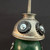 A close-up, high-angle shot of a whimsical, handcrafted found-object robot sculpture. The robot’s head is made from the silver, dome-shaped top of a vintage oil can, featuring a long vertical spout pointing upward. Two large, expressive eyes are created using black bottle caps, each accented with a silver washer and a metal Phillips-head screw in the center. 

The head sits atop the robot's body, which is a weathered and aged green vintage croquet ball with visible scratches and a rustic patina. Small metallic joints or limbs are partially visible on the sides, fastened with screws. The sculpture is set against a solid dark gray background, emphasizing the metallic textures and the upcycled folk-art aesthetic of the piece.