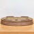 An eye-level, close-up shot of a shallow, oval-shaped ceramic bonsai pot by Jack Hoover, shown upside down on a light-colored wooden surface against a plain white background. The pot is crafted from an unglazed, earthy-brown clay with a fine, speckled, and sandy texture throughout. On its underside, four large, circular drainage holes are evenly spaced to ensure proper water flow for a bonsai tree. Four integrated, low-profile feet are positioned around the base to provide stability and lift. The outer rim of the pot features a subtle, cross-hatched or rope-like textured detail along the very edge. This handmade pot's extremely low profile and wide oval shape make it an ideal choice for forest-style bonsai arrangements or penjing.