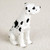 A miniature figurine of a Harlequin Great Dane dog, specifically designed as a decorative accessory for bonsai tree landscapes or fairy gardens. The small sculpture depicts the dog in an upright sitting position, facing slightly to the right. Its coat is a bright white color, accented with various irregular black patches across its body, head, and neck, characteristic of the Harlequin variety. This particular Great Dane figurine is shown with natural, uncropped ears that hang down on either side of its head. The figurine has a detailed, molded texture that mimics short fur and is placed against a neutral, off-white, slightly sandy background.
