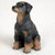 A detailed miniature figurine of a Doberman Pinscher puppy, designed as a decorative companion for a bonsai tree or miniature garden. The dog is depicted in a sitting pose, looking alertly upward and toward the left. It features the classic Doberman color pattern, with a primarily matte black body and tan (rust) markings on its muzzle, chest, and paws. This specific figurine shows the breed with natural, uncropped ears that hang down the sides of its head. The surface of the figurine has a fine, molded texture that mimics the appearance of short canine fur. It is set against a plain, off-white textured background.
