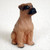 A detailed miniature figurine of a tawny Boxer dog, designed as a decorative accessory for a bonsai tree. The small figurine depicts the dog in an alert, sitting pose, looking slightly upward and to the right. It features a warm, tawny brown coat with a hand-painted black mask around the muzzle and eyes, characteristic of the Boxer breed. The figurine has a textured surface to suggest fur, small floppy ears, and black-tipped paws, all set against a soft, white textured background.