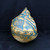 A detailed view of a 13-inch handmade crescent ceramic bonsai planter by artist John Fazzino (No. 004). The planter has a distinctive, bulbous shape that tapers into a pointed peak at the top, reminiscent of a large seed pod or an organic, weathered sculpture. Its surface is highly textured with deep, irregular ridges, impressions, and overlapping layers of clay, giving it a rugged, earth-worn appearance. 

The color palette features a warm, golden-mustard base tone that is beautifully contrasted by a vivid turquoise or blue-green glaze settled into the recessed areas and crevices. This combination creates a high-contrast, aged aesthetic that highlights the intricate hand-crafted details. This unique, one-of-a-kind bonsai pot is photographed against a solid, dark black background on a dark, horizontal surface, which emphasizes its sculptural form and rich, textured coloration. It serves as both a functional vessel for a bonsai tree and a striking piece of ceramic art.