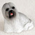 A small, intricately detailed Lhasa Apso dog figurine designed as a charming accessory for a bonsai tree display. The figurine is crafted with a realistic texture to mimic the breed's signature long, flowing coat in shades of gray and white. It features a dark face with shiny black eyes, a black nose, and a playful pink tongue peeking out. The dog is in a sitting or crouching pose, looking off-center towards the viewer's left. The entire piece is set against a neutral, light-colored background, emphasizing its delicate features and suitability for enhancing a miniature landscape or indoor garden.