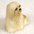 A detailed miniature figurine of a blonde Lhasa Apso dog, designed as a decorative accessory for a bonsai tree arrangement. The small sculpture captures the breed's iconic long, flowing coat with intricate, carved textures in a pale tan or cream color. The dog is depicted in a sitting position, looking off to the side with dark, expressive eyes, a small black nose, and a tiny pink tongue peeking out from its mouth. The figurine is photographed against a soft, neutral, out-of-focus background that emphasizes the fine details of the miniature's craftsmanship.
