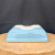 A detailed, upside-down view of a light blue glazed Yixing bonsai planter, showcased on a light-colored, circular wooden platform. This rectangular ceramic pot features inward-sloping sides and four integrated corner feet. The unglazed, off-white bottom reveals a large central drainage hole and four smaller wire holes designed for securing a bonsai tree. The pale sky-blue glaze has a glossy finish that reflects light, creating a clean and elegant aesthetic. The planter is set against a dark, solid-colored backdrop that makes the light blue color pop.