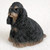 A detailed miniature figurine of a Black and Tan Cocker Spaniel, designed as a decorative accessory for bonsai tree displays. The small statue depicts the dog in a sitting pose, looking slightly to the side with a gentle and soulful expression. The figurine features a highly textured surface that realistically mimics the breed's characteristic long, wavy coat. Its primary color is a deep, rich black, accented with classic tan markings on its muzzle, as small spots above its dark eyes, and along the bottom edge to represent its paws. This charming miniature dog is crafted to add a touch of personality and life to a bonsai arrangement, fairy garden, or any small-scale indoor landscape.