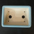 A top-down view of a 7-inch glazed rectangle Yixing ceramic bonsai pot (No. 2327C), highlighting its functional design for bonsai cultivation. The pot features a pale blue glazed rim that contrasts with its unglazed, tan-colored interior. 

Centered on the bottom are two large, round drainage holes to prevent overwatering. Four smaller holes are positioned near the corners, specifically designed for anchoring wire to hold a bonsai tree securely in place. The pot's rectangular form has soft, rounded corners and sides that slope inward towards the base, all set against a dark black background to emphasize its clean lines and subtle color.