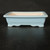 A side-profile view of a 7-inch rectangular Yixing ceramic bonsai pot with a soft, light-blue glaze. The pot features a classic rectangular shape with a slightly flared rim and four decorative, cloud-shaped feet at the corners. The exterior is finished with a smooth, glossy, powder-blue glaze that contrasts with the unglazed interior, which reveals the natural, light-cream color of the fired clay. This porous interior is characteristic of traditional Yixing pottery, which is highly valued for its ability to promote healthy root growth in bonsai trees. The pot is centered in the frame against a minimalist dark black background, resting on a dark surface that reflects a hint of the blue glaze, emphasizing its elegant form and serene color.