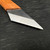 A detailed close-up of a professional-grade right-hand bonsai grafting knife, model GFN-03, resting on a dark, scratched work surface. The image highlights the tool's sharp, single-bevel steel blade, which features a highly polished, ground edge that contrasts with the darker, matte finish of the upper blade. The blade tapers to an exceptionally fine, sharp point, essential for making the precise, clean cuts required for successful bonsai grafting. The blade is set into a simple, straight handle made of natural, light-colored wood with a visible grain, providing a comfortable and traditional grip for delicate horticultural work. The dark, textured background further emphasizes the knife's sharpness and its utility as a precision tool for bonsai enthusiasts.