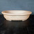 An oval-shaped, unglazed Yixing clay bonsai pot, approximately 16 inches long, in a light tan or buff color. The pot has a shallow, elegant profile with a wide, flat rim and subtle horizontal ribbing around its exterior. Small feet at the base provide elevation for drainage. The pot is positioned on a dark, textured surface against a charcoal-gray background, highlighting its smooth, matte, earthy texture and minimalist design.