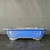 A large, 18-inch rectangular Yixing bonsai pot is shown in a profile view, showcasing its deep cobalt blue glaze with a subtle mottled texture. The pot features a classic design with a wide rim that flares out slightly at the corners. On its long side, there is a recessed decorative rectangular panel that adds a structured, architectural feel to the piece. The pot stands on four short, sturdy block-style feet, elevating the base. It is resting on a dark, wood-textured surface against a neutral, dark gray fabric backdrop, which makes the vibrant blue color of the ceramic stand out prominently.