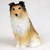 A detailed miniature figurine of a Shetland Sheepdog, or Sheltie, in a sitting pose against a plain, off-white background. The small dog statue features a realistic sable coat pattern, with golden-tan fur on its body and a large, textured white ruff around its neck and chest that extends down its front legs. The top of the head and the tips of its semi-erect ears are shaded with dark brown and black, highlighting its attentive expression. Its small black eyes and nose are finely detailed, adding to its lifelike appearance. This tiny figurine is designed as a bonsai tree accessory, intended to bring a charming, realistic element to miniature garden displays or indoor plant arrangements.