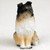 A detailed miniature figurine of a tri-color Shetland Sheepdog, commonly known as a Sheltie, designed as a decorative accessory for a bonsai tree. The small dog figurine is depicted in a sitting position, looking alertly toward the right. Its tri-color coat is meticulously sculpted to show the texture of long, thick fur, featuring black on the head and ears, rich tan markings on the face and legs, and a bright white chest ruff and front paws. The Sheltie has characteristic semi-erect ears and dark, expressive eyes, capturing the lifelike essence of the breed in a miniature scale. The figurine is set against a neutral, off-white background, highlighting its intricate paintwork and realistic features.